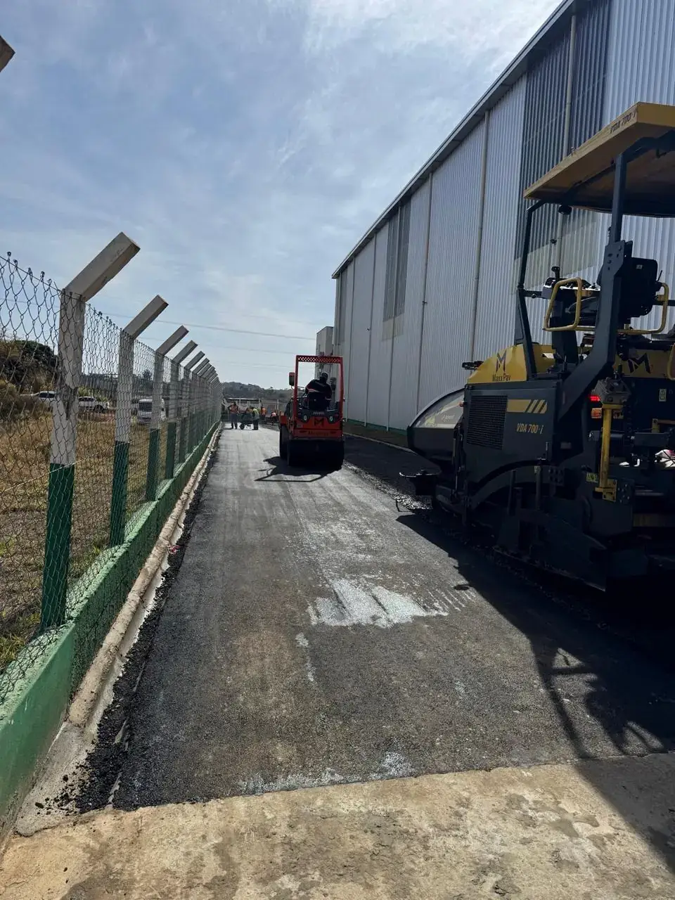 Base com ligante asfáltico aplicado e vibroacabadora ao fundo, pronta para receber a capa de CBUQ