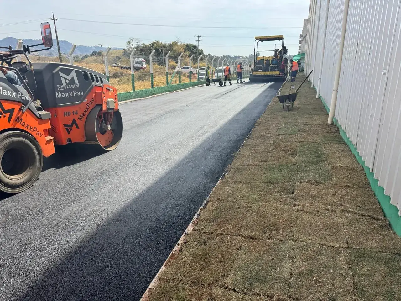 Rolo liso dando acabamento à capa asfáltica enquanto a vibroacabadora avança ao fundo